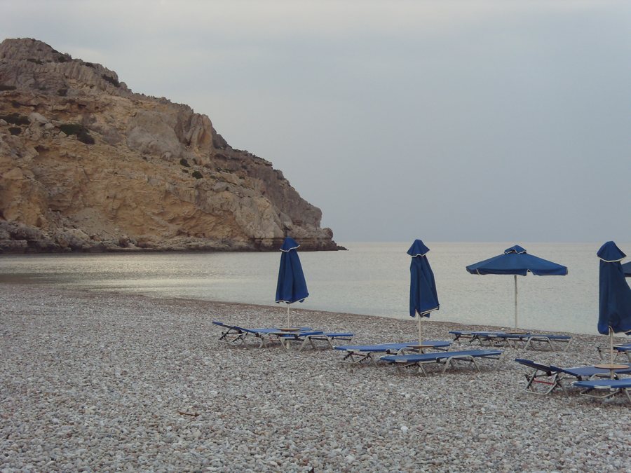 Kiesstrand auf Rhodos mit Sonnenschirmen vor einem Felsufer.